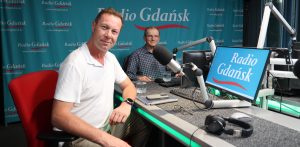 Na zdjęciu dwóch mężczyzn siedzi w studiu nagraniowym przed mikrofonami. Jeden ma na sobie biały tshirt, a drugi koszulę w kratę. Na stole stoi monitor, klawiatura i leżą okulary, telefon i tablet, a także słuchawki radiowe.