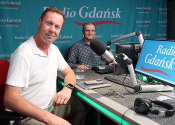 Na zdjęciu dwóch mężczyzn siedzi w studiu nagraniowym przed mikrofonami. Jeden ma na sobie biały tshirt, a drugi koszulę w kratę. Na stole stoi monitor, klawiatura i leżą okulary, telefon i tablet, a także słuchawki radiowe.