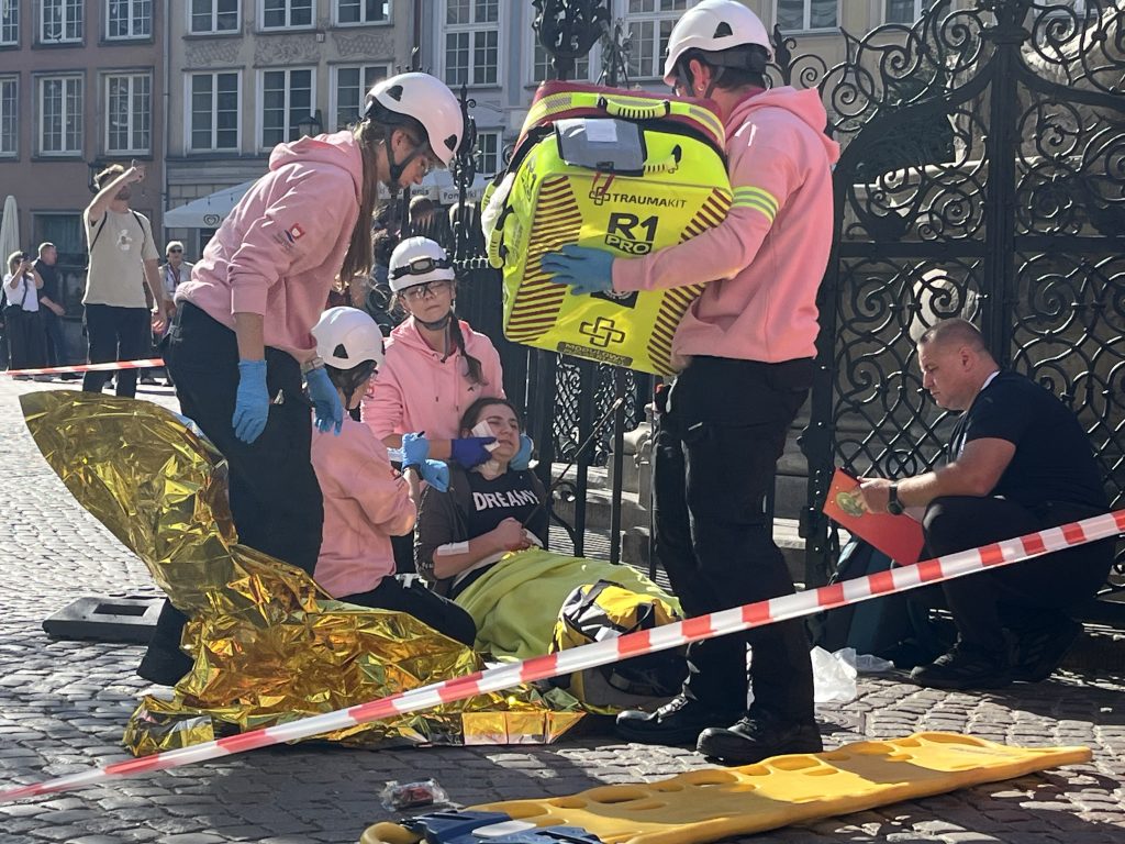 Drużyna ratownicza ZHP w różowych bluzach i kaskach zabezpiecza poszkodowaną osobę leżącą przy ozdobnej bramie, używając torby medycznej R1 Pro oraz folii NRC. Przebieg symulacji jest oceniany przez sędziego siedzącego obok z podkładką i formularzem ocen