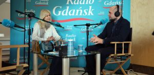 Joanna Łapińska, dyrektor artystyczna Festiwalu Polskich Filmów Fabularnych w Gdyni, oraz dziennikarz Kuba Kaługa w słuchawkach rozmawiają przy stole przed mikrofonami w studiu Radia Gdańsk na tle błękitnej ścianki z logo stacji