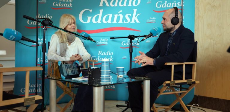 Joanna Łapińska, dyrektor artystyczna Festiwalu Polskich Filmów Fabularnych w Gdyni, oraz dziennikarz Kuba Kaługa w słuchawkach rozmawiają przy stole przed mikrofonami w studiu Radia Gdańsk na tle błękitnej ścianki z logo stacji