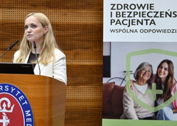 Na zdjęciu kobieta przy mikronofie na scenie. Za nią rollup z napisem "Zdrowie i bezpieczeństwo pacjenta".