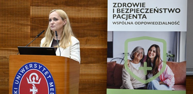 Na zdjęciu kobieta przy mikronofie na scenie. Za nią rollup z napisem "Zdrowie i bezpieczeństwo pacjenta".