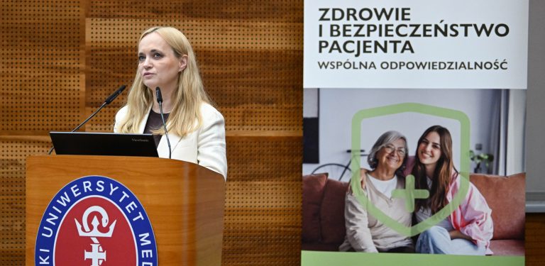 Na zdjęciu kobieta przy mikronofie na scenie. Za nią rollup z napisem "Zdrowie i bezpieczeństwo pacjenta".