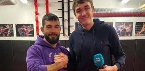 Zawodnik MMA Kacper Formela w fioletowej bluzie i dziennikarz Radia Gdańsk Karol Pius trzymający mikrofon stacji, uśmiechają się i podają sobie ręce na tle sali treningowej.