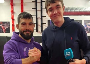 Zawodnik MMA Kacper Formela w fioletowej bluzie i dziennikarz Radia Gdańsk Karol Pius trzymający mikrofon stacji, uśmiechają się i podają sobie ręce na tle sali treningowej.