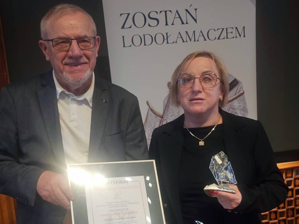 Mecenas Katarzyna Heba trzymająca statuetkę i certyfikat tytułu honorowego "Lodołamacz Specjalny 2026" w towarzystwie mężczyzny, na tle ścianki z hasłem "Zostań Lodołamaczem"