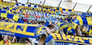 Żółto-niebieskie szaliki klubu Arka Gdynia w uniesionych ponad głowy rękach kibiców stojących na trybunach stadionu. Zbliżenie na jeden z sektorów