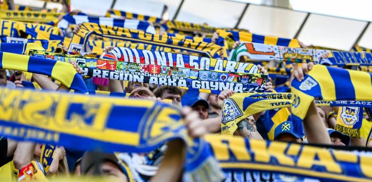 Żółto-niebieskie szaliki klubu Arka Gdynia w uniesionych ponad głowy rękach kibiców stojących na trybunach stadionu. Zbliżenie na jeden z sektorów