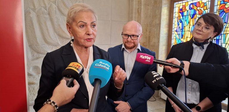 Prezydent Słupska Krystyna Danilecka-Wojewódzka wypowiada się do mikrofonów dziennikarzy podczas konferencji prasowej. W tle stoją miejscy urzędnicy oraz widoczne jest witrażowe okno