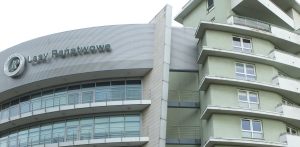Fragment wielopiętrowego budynku biurowo-mieszkalnego z widocznym na zaokrąglonej elewacji logotypem oraz napisem "Lasy Państwowe". Do sądu wraca sprawa byłych przedstawicieli kierownictwa instytucji na Pomorzu, oskarżonych o przekroczenie uprawnień i próbę sprzedaży lokali mieszkalnych w Gdyni, co mogło narazić Lasy Państwowe na szkodę majątkową