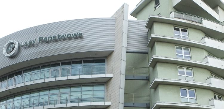 Fragment wielopiętrowego budynku biurowo-mieszkalnego z widocznym na zaokrąglonej elewacji logotypem oraz napisem "Lasy Państwowe". Do sądu wraca sprawa byłych przedstawicieli kierownictwa instytucji na Pomorzu, oskarżonych o przekroczenie uprawnień i próbę sprzedaży lokali mieszkalnych w Gdyni, co mogło narazić Lasy Państwowe na szkodę majątkową