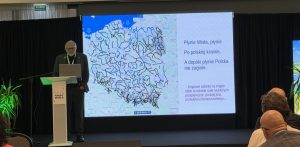 Prelegent prof. Szymon Malinowski stoi przy mównicy podczas konferencji "Łączy nas Wisła" na tle wielkiego ekranu z mapą hydrologiczną rzek w Polsce. Obok mapy widnieje tekst piosenki "Płynie Wisła, płynie" oraz dopisek o odcinkach rzek z przepływem poniżej normy. Na pierwszym planie widać słuchających uczestników wydarzenia