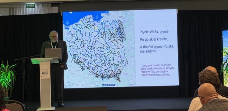 Prelegent prof. Szymon Malinowski stoi przy mównicy podczas konferencji "Łączy nas Wisła" na tle wielkiego ekranu z mapą hydrologiczną rzek w Polsce. Obok mapy widnieje tekst piosenki "Płynie Wisła, płynie" oraz dopisek o odcinkach rzek z przepływem poniżej normy. Na pierwszym planie widać słuchających uczestników wydarzenia