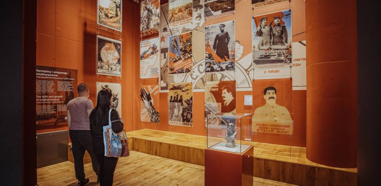Dwoje zwiedzających ogląda ekspozycję w Muzeum II Wojny Światowej w Gdańsku. Na pomarańczowej ścianie wiszą radzieckie plakaty propagandowe przedstawiające m.in. Józefa Stalina, a na podeście w szklanej gablocie stoi niewielkie popiersie. Placówka, we współpracy z kołem PSONI, przygotowała nowe ścieżki zwiedzania dostosowane do potrzeb osób z niepełnosprawnością intelektualną, w tym materiały w tekście łatwym do zrozumienia (ETR) oraz audioprzewodnik czytany przez Annę Dymną