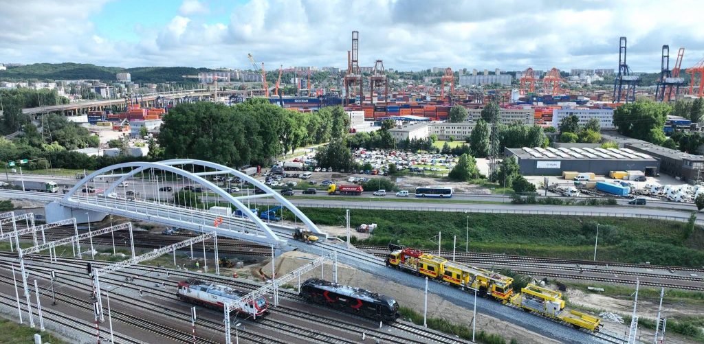 Widok z lotu ptaka na nowy, biały łukowy wiadukt kolejowy w Gdyni. Pod konstrukcją i obok niej biegną liczne tory kolejowe, po których poruszają się lokomotywy oraz żółte maszyny torowe. W tle widać ruchliwą ulicę Janka Wiśniewskiego oraz rozległą panoramę portu morskiego z suwnicami kontenerowymi