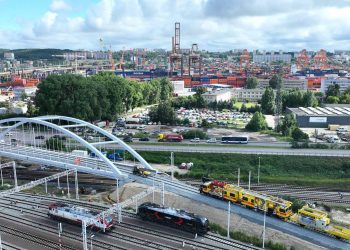 Widok z lotu ptaka na nowy, biały łukowy wiadukt kolejowy w Gdyni. Pod konstrukcją i obok niej biegną liczne tory kolejowe, po których poruszają się lokomotywy oraz żółte maszyny torowe. W tle widać ruchliwą ulicę Janka Wiśniewskiego oraz rozległą panoramę portu morskiego z suwnicami kontenerowymi
