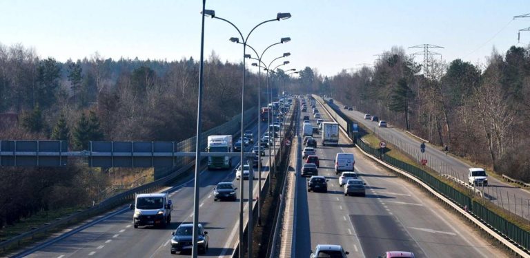 Widok z wiaduktu na wielopasmową drogę ekspresową (Obwodnicę Trójmiasta) w słoneczny dzień. Pomiędzy jezdniami biegnie rząd wysokich latarni ulicznych. Po pasach poruszają się samochody osobowe, dostawcze i ciężarowe, a na jednej z nitek tworzy się zator. Po obu stronach trasy rośnie las.