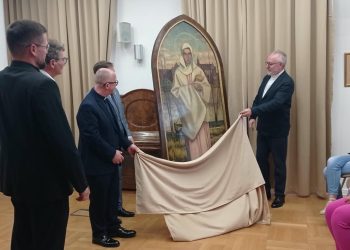 Mężczyźni w garniturach i sutannach uroczyście zdejmują beżową płachtę materiału, odsłaniając duży, zwieńczony łukiem obraz przedstawiający Matkę Boską na tle zamku. Z prawej strony ceremonię obserwują siedzące kobiety. To Uroczyste odsłonięcie odnalezionego po ponad 80 latach obrazu Matki Boskiej Malborskiej z 1942 roku autorstwa Hansa Franke