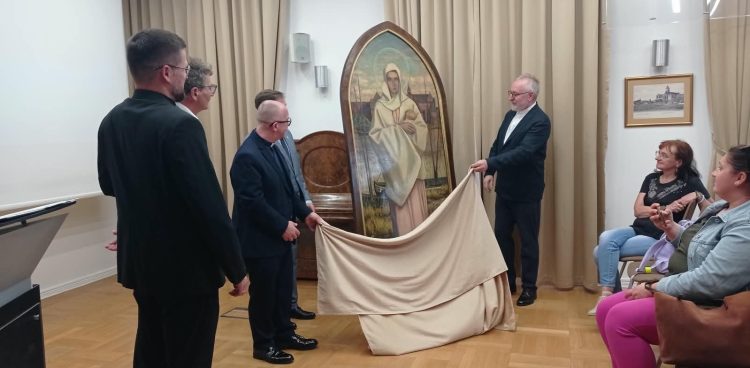 Mężczyźni w garniturach i sutannach uroczyście zdejmują beżową płachtę materiału, odsłaniając duży, zwieńczony łukiem obraz przedstawiający Matkę Boską na tle zamku. Z prawej strony ceremonię obserwują siedzące kobiety. To Uroczyste odsłonięcie odnalezionego po ponad 80 latach obrazu Matki Boskiej Malborskiej z 1942 roku autorstwa Hansa Franke