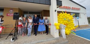 Uroczyste przecięcie czerwonej wstęgi przed nowo wybudowanym Żłobkiem Gminnym "Promyczek" w Głobinie (gmina Redzikowo). Dorośli oraz małe dzieci wspólnie trzymają nożyczki, a obok wejścia stoi duża dekoracja ze słoneczkiem z żółtych balonów. Dzięki rządowemu wsparciu w regionie powstają kolejne żłobki, które zaczynają konkurować