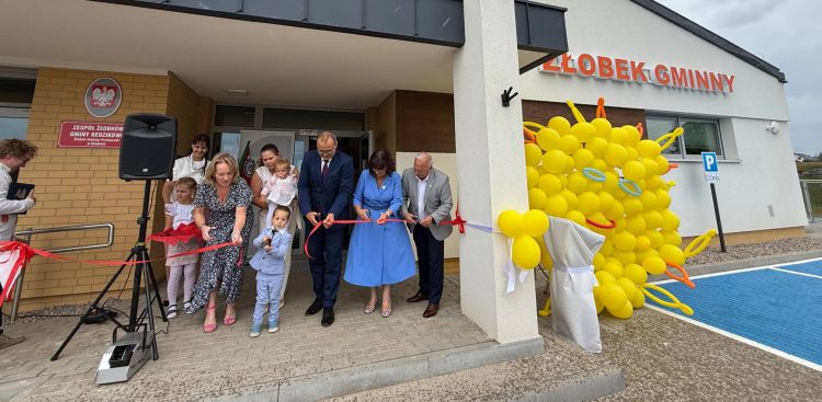 Uroczyste przecięcie czerwonej wstęgi przed nowo wybudowanym Żłobkiem Gminnym "Promyczek" w Głobinie (gmina Redzikowo). Dorośli oraz małe dzieci wspólnie trzymają nożyczki, a obok wejścia stoi duża dekoracja ze słoneczkiem z żółtych balonów. Dzięki rządowemu wsparciu w regionie powstają kolejne żłobki, które zaczynają konkurować
