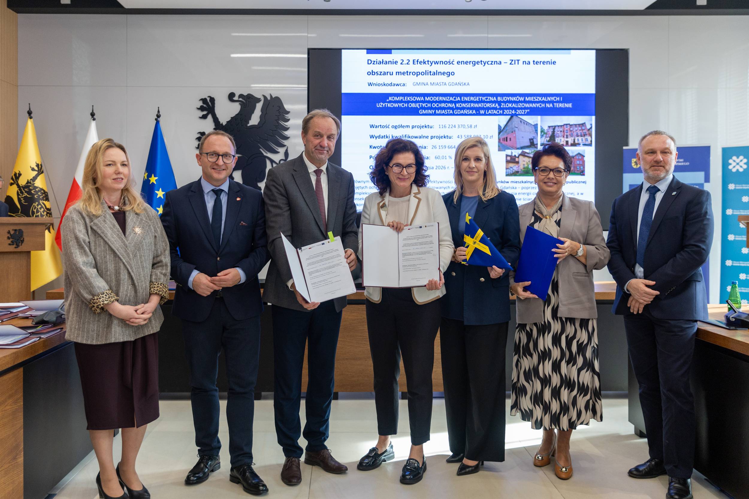 Grupa pomorskich samorządowców stoi w sali konferencyjnej. W centrum prezydent Gdańska Aleksandra Dulkiewicz oraz marszałek województwa Mieczysław Struk prezentują otwarty, podpisany dokument. Obok nich stoją m.in. wiceprezydent Gdańska Emilia Lodzińska, wicemarszałkowie oraz samorządowcy. W tle znajduje się ekran z prezentacją projektu termomodernizacji oraz flagi
