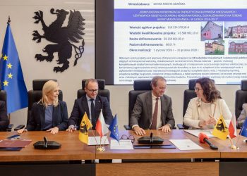 Przedstawiciele pomorskich samorządów, w tym marszałek Mieczysław Struk oraz zastępca prezydenta Gdańska Emilia Lodzińska, siedzą przy stole konferencyjnym podczas podpisywania umów. W tle widoczne są flagi oraz ekran z prezentacją projektu termomodernizacji budynków w Gdańsku