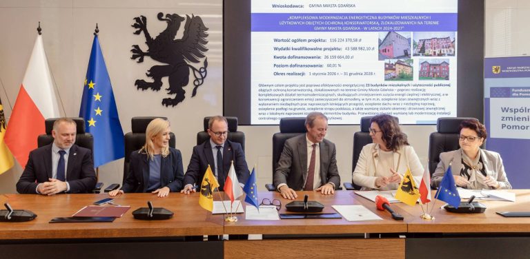 Przedstawiciele pomorskich samorządów, w tym marszałek Mieczysław Struk oraz zastępca prezydenta Gdańska Emilia Lodzińska, siedzą przy stole konferencyjnym podczas podpisywania umów. W tle widoczne są flagi oraz ekran z prezentacją projektu termomodernizacji budynków w Gdańsku