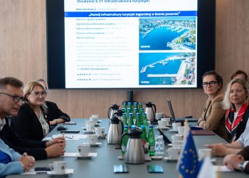 Uczestnicy spotkania przy stole konferencyjnym, w tle ekran z prezentacją projektu rozwoju przystani w Jastarni i Kuźnicy