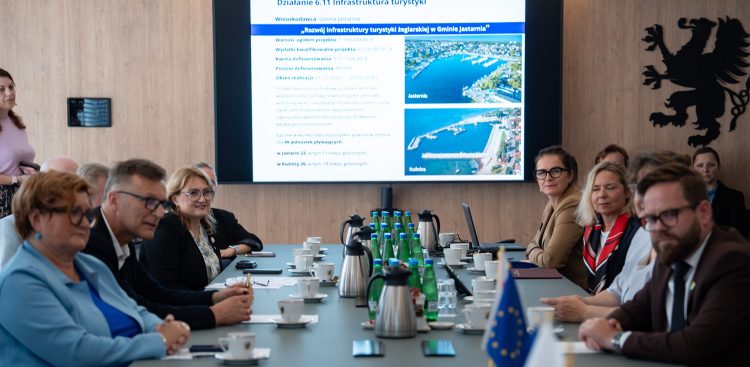 Uczestnicy spotkania przy stole konferencyjnym, w tle ekran z prezentacją projektu rozwoju przystani w Jastarni i Kuźnicy