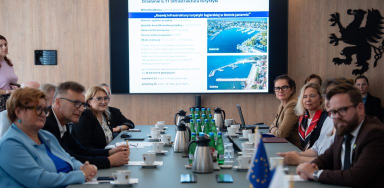 Uczestnicy spotkania przy stole konferencyjnym, w tle ekran z prezentacją projektu rozwoju przystani w Jastarni i Kuźnicy