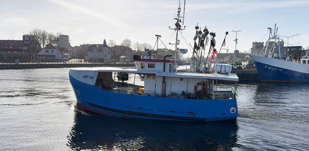 Niebiesko-biały kuter rybacki o oznaczeniu UST 130 płynący kanałem portowym w Ustce; w tle widoczne nabrzeże, zabudowa portowa oraz zacumowany większy kuter AGAT UST-113. Wzrost populacji śledzia i szprota w Morzu Bałtyckim oraz planowane przez Komisję Europejską podwyższenie limitów połowowych dają armatorom i rybakom nadzieję na poprawę trudnej sytuacji w branży