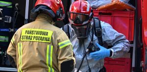 Dwóch strażaków Państwowej Straży Pożarnej przy wozie bojowym. Jeden z nich jest w specjalistycznym kombinezonie chemicznym, masce aparatu oddechowego i czerwonym hełmie. Teren placówki opiekuńczo-wychowawczej przy ulicy Mazurskiej w Elblągu, 29 bm. Policja ewakuowała 30 dzieci, w jednym z pokoi tej placówki wybuchł pożar