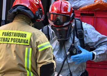 Dwóch strażaków Państwowej Straży Pożarnej przy wozie bojowym. Jeden z nich jest w specjalistycznym kombinezonie chemicznym, masce aparatu oddechowego i czerwonym hełmie. Teren placówki opiekuńczo-wychowawczej przy ulicy Mazurskiej w Elblągu, 29 bm. Policja ewakuowała 30 dzieci, w jednym z pokoi tej placówki wybuchł pożar