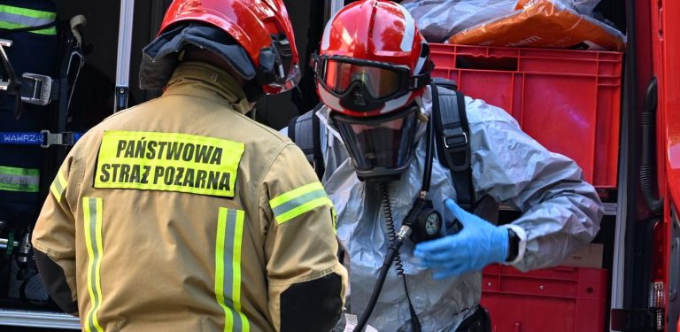 Dwóch strażaków Państwowej Straży Pożarnej przy wozie bojowym. Jeden z nich jest w specjalistycznym kombinezonie chemicznym, masce aparatu oddechowego i czerwonym hełmie. Teren placówki opiekuńczo-wychowawczej przy ulicy Mazurskiej w Elblągu, 29 bm. Policja ewakuowała 30 dzieci, w jednym z pokoi tej placówki wybuchł pożar