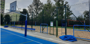 Fragment niebieskiego boiska wielofunkcyjnego z koszem do koszykówki, rzędem niebieskich krzesełek oraz ogrodzonym, piaszczystym boiskiem do siatkówki plażowej z siatkami. W tle widoczne drzewa i blok mieszkalny. To nowy przyszkolny kompleks sportowy, największy w Gdyni