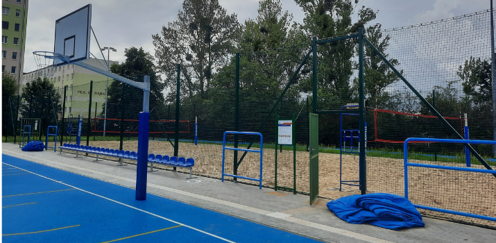 Fragment niebieskiego boiska wielofunkcyjnego z koszem do koszykówki, rzędem niebieskich krzesełek oraz ogrodzonym, piaszczystym boiskiem do siatkówki plażowej z siatkami. W tle widoczne drzewa i blok mieszkalny. To nowy przyszkolny kompleks sportowy, największy w Gdyni
