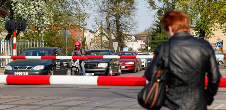 Zamknięte rogatki na przejeździe kolejowym w Gdańsku. Czekają samochody i motocyklista. Zdjęcie ilustruje temat modernizacji trasy z Lipusza do Bytowa. PKP PLK planuje tam montaż rogatek na połowie przejazdów. Mieszkańcy mają czas do końca września na składanie wniosków dotyczących dostępu do nich