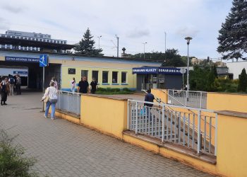 Plac i wejście do tunelu podziemnego przy przystanku SKM Gdynia Wzgórze Świętego Maksymiliana. Po prawej stronie schody w dół otoczone żółtym murkiem i barierkami, w tle parterowy budynek stacji, punkty usługowe oraz przechodnie na chodniku