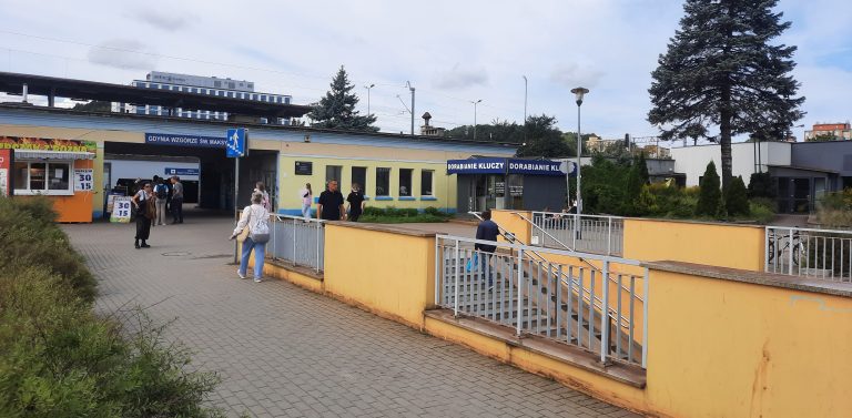 Plac i wejście do tunelu podziemnego przy przystanku SKM Gdynia Wzgórze Świętego Maksymiliana. Po prawej stronie schody w dół otoczone żółtym murkiem i barierkami, w tle parterowy budynek stacji, punkty usługowe oraz przechodnie na chodniku