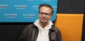 Mężczyzna w okularach i z zarostem, ubrany w jasną bluzę z kapturem i ciemną skórzaną kurtkę – Radosław Paczocha, zastępca dyrektora Teatru Wybrzeże. Siedzi w studiu radiowym na tle ścianki z logo Radia Gdańsk i paneli akustycznych