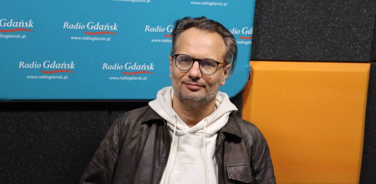 Mężczyzna w okularach i z zarostem, ubrany w jasną bluzę z kapturem i ciemną skórzaną kurtkę – Radosław Paczocha, zastępca dyrektora Teatru Wybrzeże. Siedzi w studiu radiowym na tle ścianki z logo Radia Gdańsk i paneli akustycznych