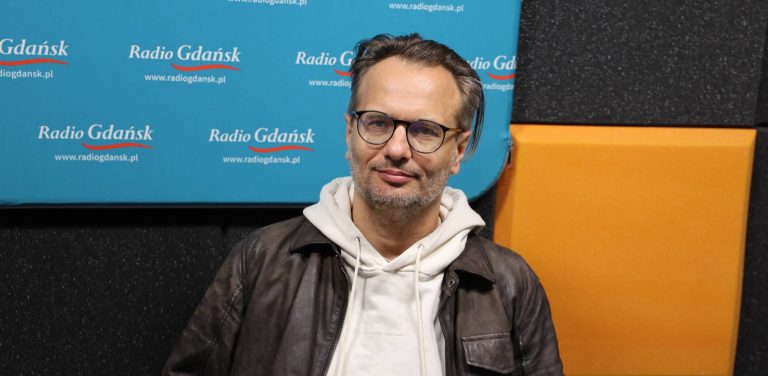 Mężczyzna w okularach i z zarostem, ubrany w jasną bluzę z kapturem i ciemną skórzaną kurtkę – Radosław Paczocha, zastępca dyrektora Teatru Wybrzeże. Siedzi w studiu radiowym na tle ścianki z logo Radia Gdańsk i paneli akustycznych