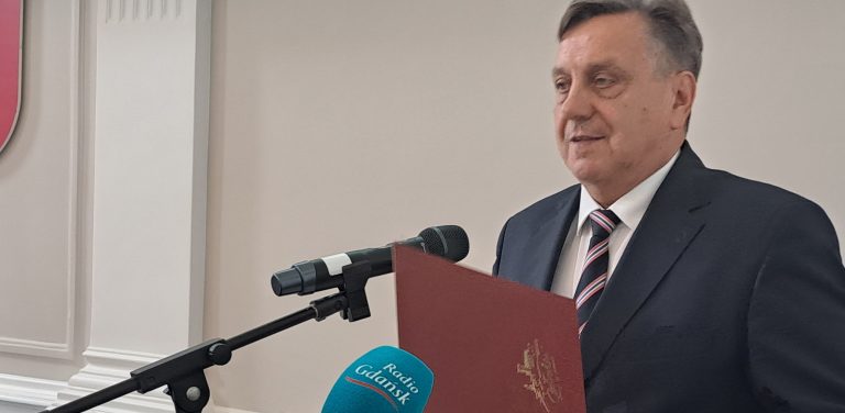 Burmistrz Człuchowa Ryszard Szybajło w ciemnym garniturze i krawacie, stojący przed mikrofonem i trzymający bordową teczkę. Decyzją radnych miejskich jego miesięczne wynagrodzenie wzrośnie o ponad 600 zł brutto, osiągając kwotę ponad 21 tys. zł brutto