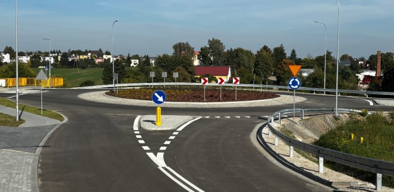 Nowo wybudowane rondo drogowe obwodnicy Skórcza z gładką nawierzchnią asfaltową, białym oznakowaniem poziomym, znakami drogowymi i barierami ochronnymi. W centralnej części ronda znajduje się wyspa z nasadzeniami, a w tle widać zieleń oraz zabudowę jednorodzinną pod pogodnym niebem.