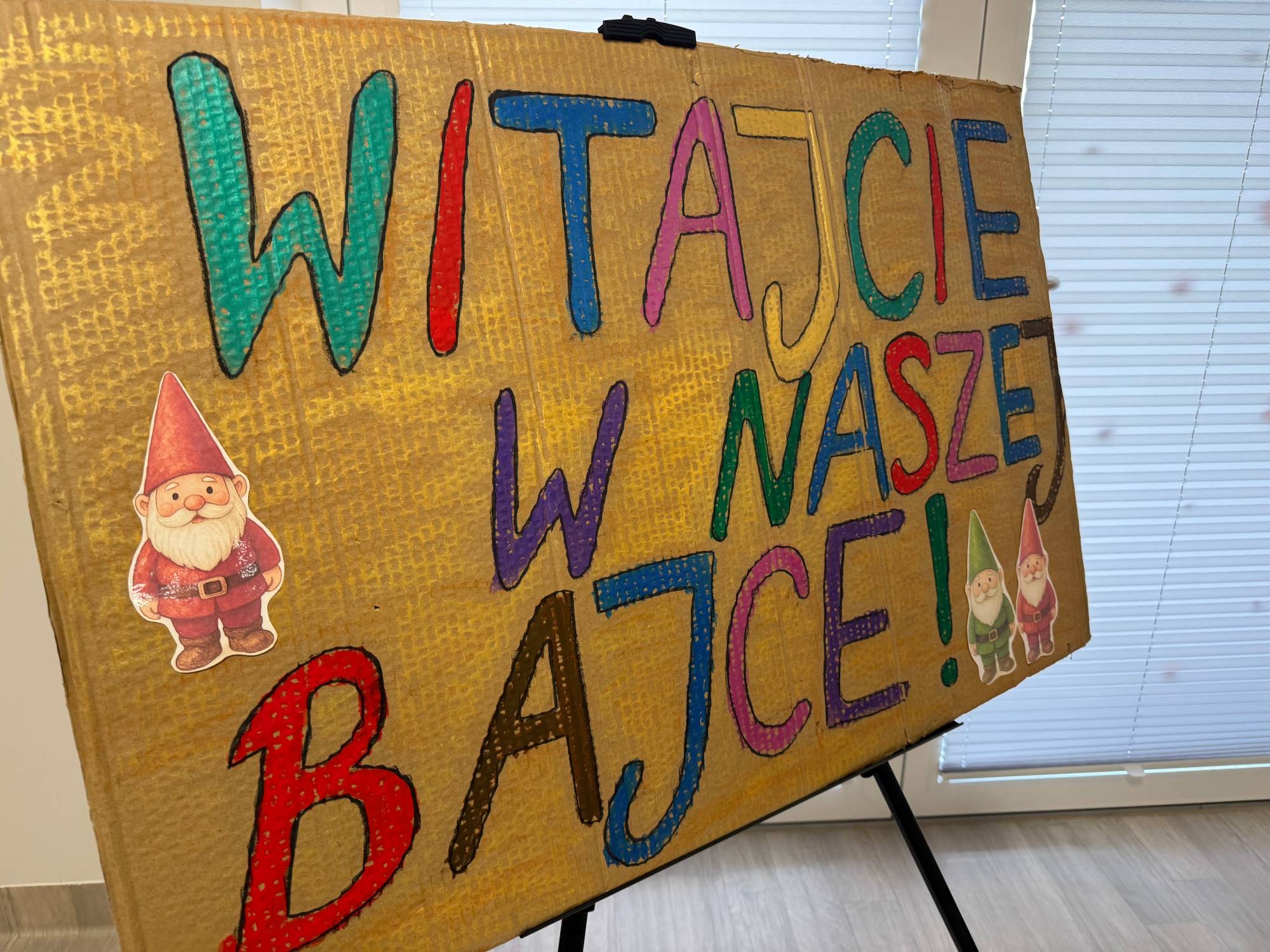 Wnętrze żłobka. Ręcznie wykonany, tekturowy plakat na sztaludze z dużym, kolorowym napisem „WITAJCIE W NASZEJ BAJCE!”. Na rogach tablicy doklejone są wycięte ilustracje uśmiechniętych krasnoludków