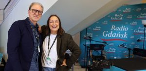 Uśmiechnięci dziennikarze Radia Gdańsk, Tomasz Żółtowski i Magda Manasterska, pozują w festiwalowym studiu 51. FPFF na tle niebieskiej ścianki z logo Radia Gdańsk. Na pierwszym planie widoczna jest kamera wideo oraz mikrofony radiowe