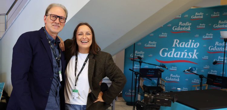 Uśmiechnięci dziennikarze Radia Gdańsk, Tomasz Żółtowski i Magda Manasterska, pozują w festiwalowym studiu 51. FPFF na tle niebieskiej ścianki z logo Radia Gdańsk. Na pierwszym planie widoczna jest kamera wideo oraz mikrofony radiowe