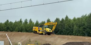 Żółta koparka gąsienicowa marki Komatsu oraz walec drogowy stojące na piaszczystym nasypie kolejowym na tle ściany lasu. U góry widoczne przewody sieci trakcyjnej. Maszyny budowlane na nasypie ziemnym w rejonie Głuszyna podczas prac przy budowie drugiego toru linii kolejowej nr 202. W tym rejonie doszło do tragicznego wypadku, w którym pod gąsienicami koparki zginęła 45-letnia pracownica